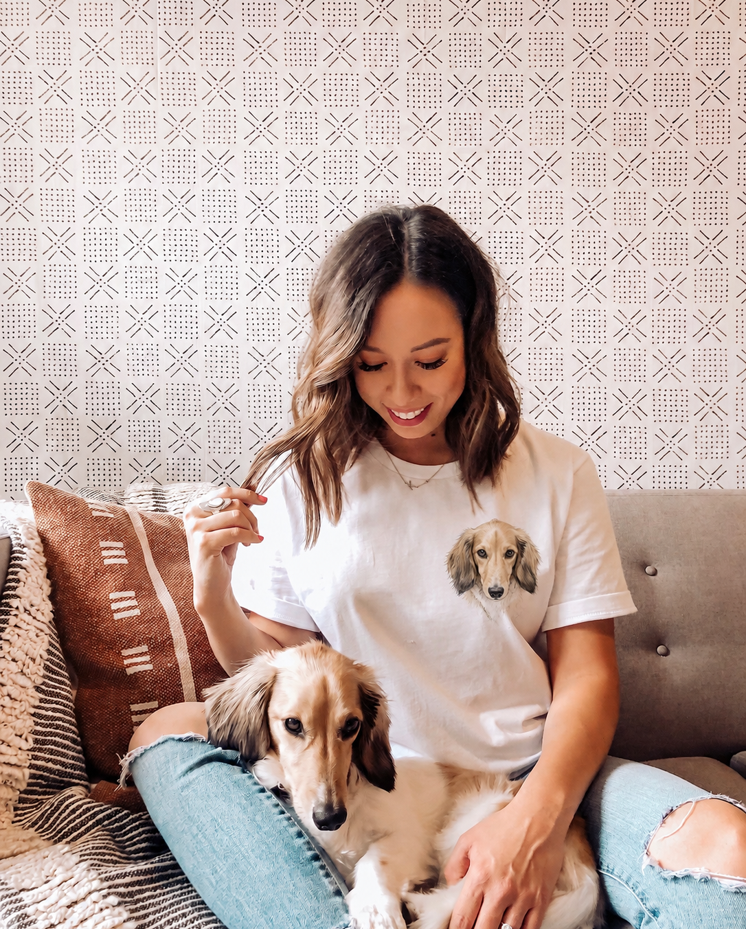 A woman sitting on a gray tufted couch with a dachshund, wearing a white graphic t-shirt with a printed dachshund face on the chest.