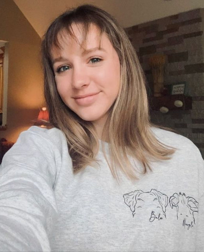 A woman taking a selfie in a home setting, wearing a light gray heather crewneck sweatshirt with a black line art graphic of two horses and the text 'Bila Rug' on the front.