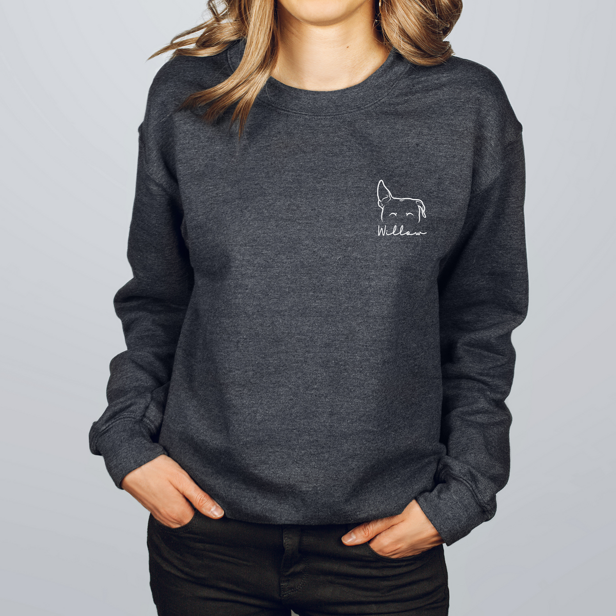 A woman wearing a dark heather gray crewneck sweatshirt with a small white embroidered dog outline and the brand name 'Wildawn' on the left chest, paired with black pants against a light gray studio background.