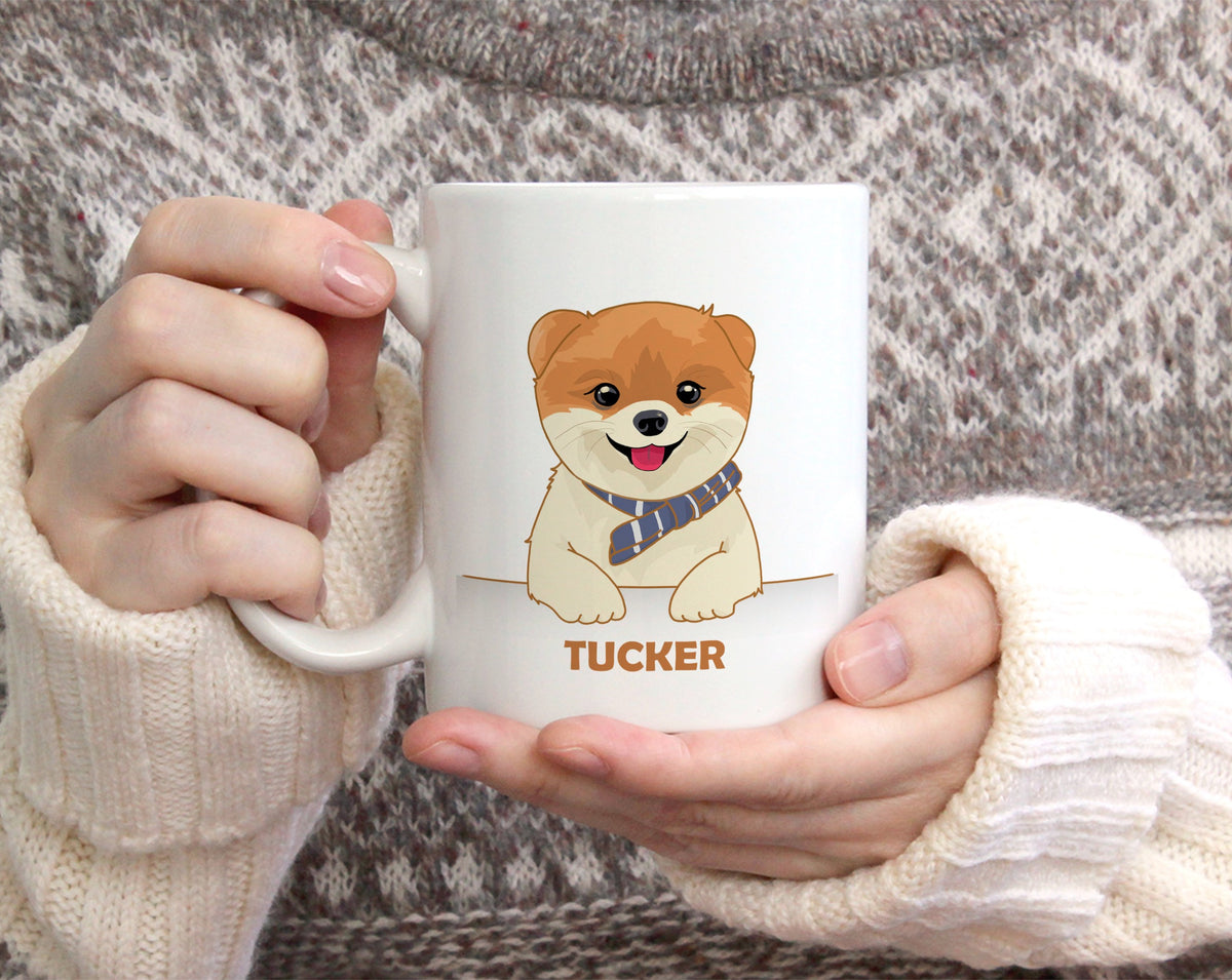 A person wearing a gray and beige knit sweater holds a white ceramic mug with a cartoon Shiba Inu dog illustration and the name 'TUCKER' printed below.