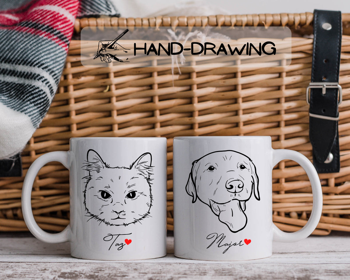 Two white ceramic mugs displayed in front of a woven wicker basket. The left mug features a black line art drawing of a cat with the name 'Tos' and a small red heart.