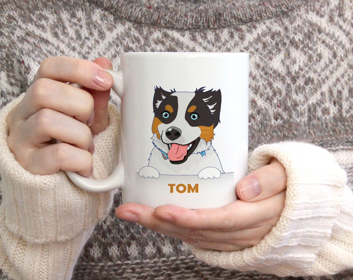 A person holding a white ceramic mug with a cartoon dog illustration and the name 'TOM' printed on it, wearing a cream knit sweater.