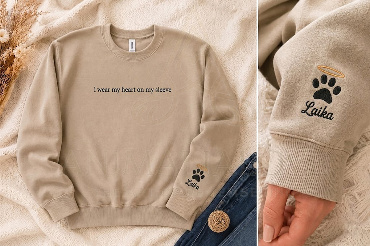 A beige crewneck sweatshirt laid flat on a soft cream fabric background, with a close-up inset showing the left sleeve embroidery. The sweatshirt has black text reading 'i wear my heart on my sleeve' across the chest and a paw print with the name 'Laika' embroidered on the sleeve.