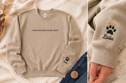 A beige crewneck sweatshirt laid flat on a soft cream fabric background, with a close-up inset showing the left sleeve embroidery. The sweatshirt has black text reading 'i wear my heart on my sleeve' across the chest and a paw print with the name 'Laika' embroidered on the sleeve.