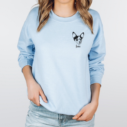 A woman wearing a light blue crewneck sweatshirt with a black graphic of a dog's face and the word 'Juno' on the left chest, paired with light-wash jeans against a light gray background.