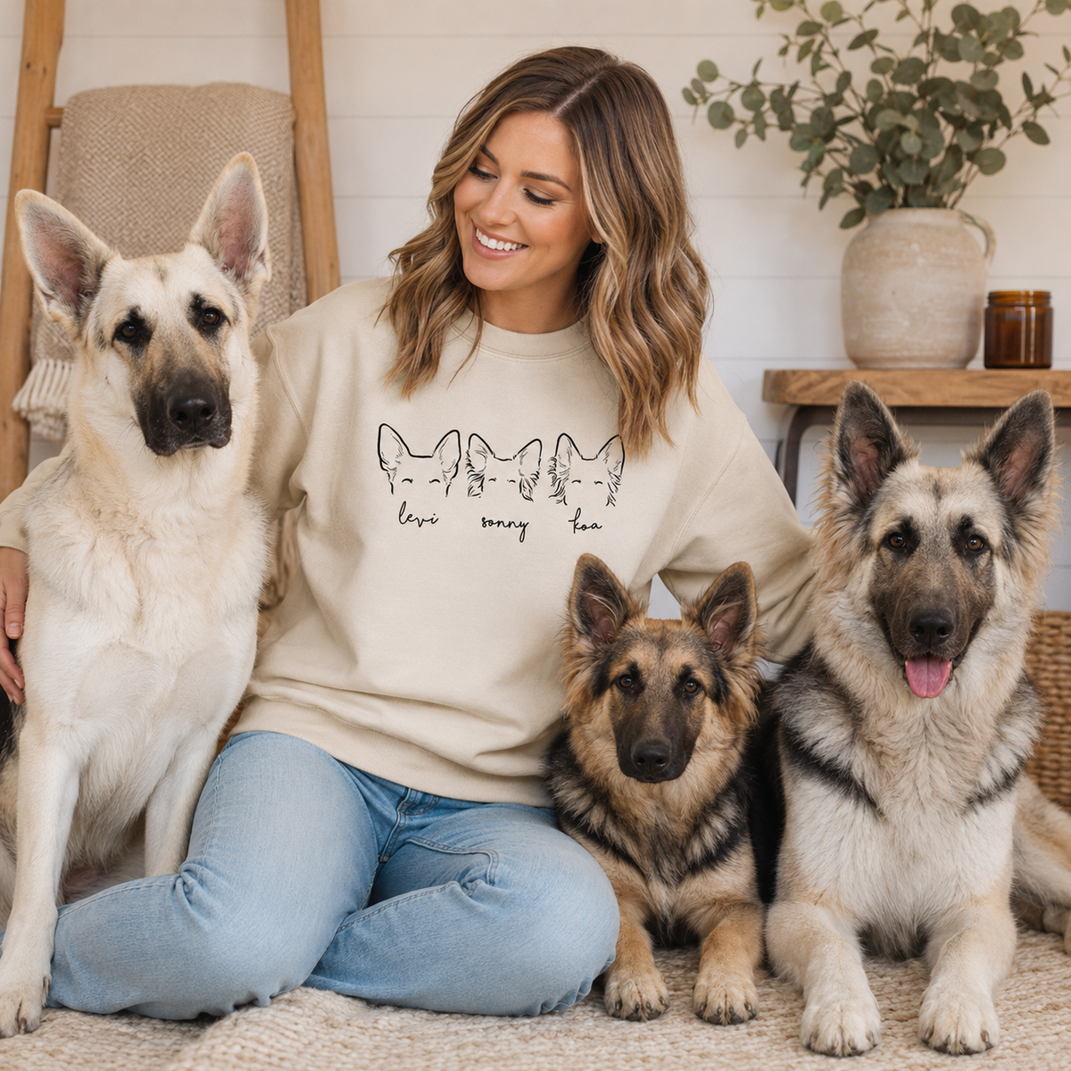 A woman sitting on a rug with three dogs, wearing a beige crewneck sweatshirt with a black graphic of four cats and the text 'love every cat'.