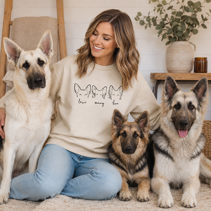 A woman sitting on a rug with three dogs, wearing a beige crewneck sweatshirt with a black graphic of four cats and the text 'love every cat'.