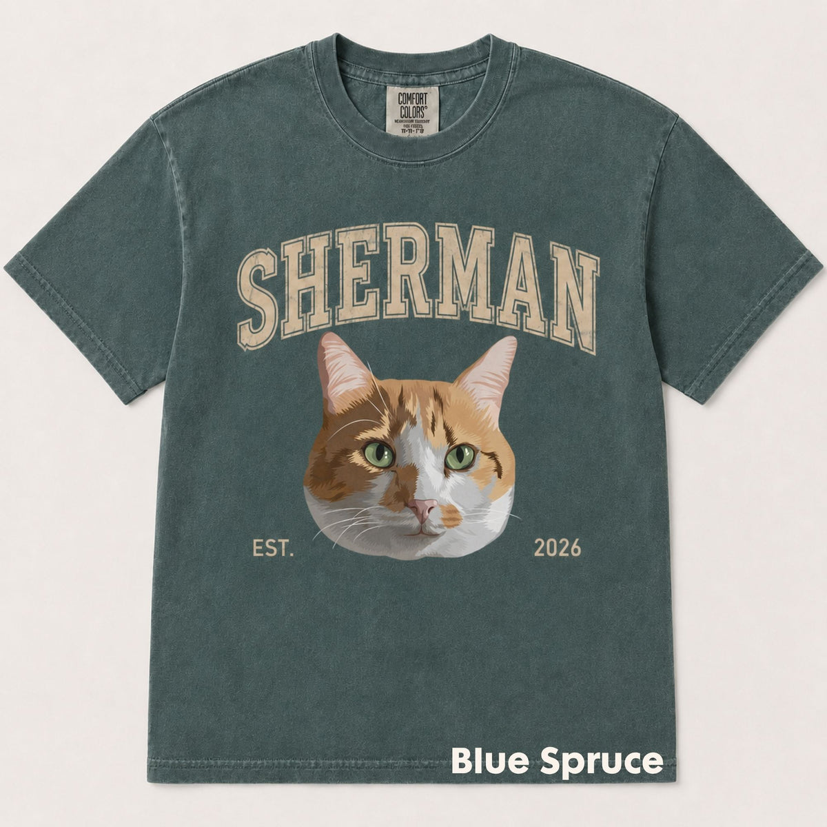 A washed slate blue-green t-shirt featuring a graphic of an orange and white cat with green eyes, with the word 'SHERMAN' above and 'EST. 2026' below.