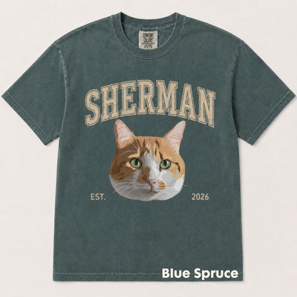 A washed slate blue-green t-shirt featuring a graphic of an orange and white cat with green eyes, with the word 'SHERMAN' above and 'EST. 2026' below.