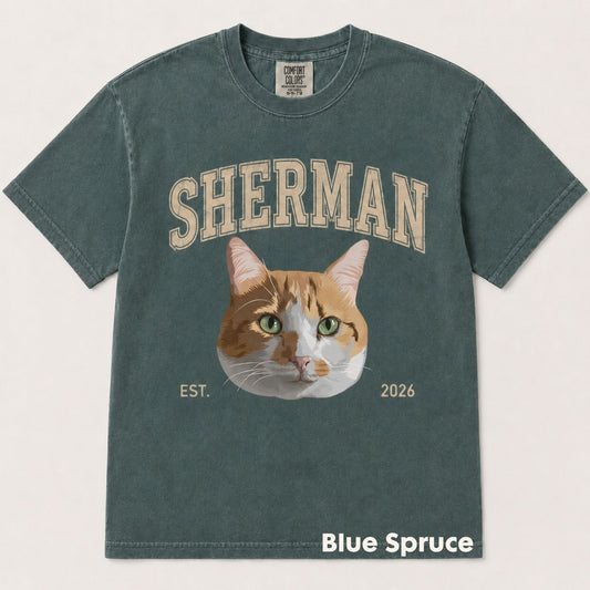 A washed slate blue-green t-shirt featuring a graphic of an orange and white cat with green eyes, with the word 'SHERMAN' above and 'EST. 2026' below.