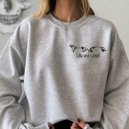 A person wearing a ash color crewneck sweatshirt with a black graphic of two dogs and the text 'Lily and Licent' on the chest, shown in a styled indoor setting.