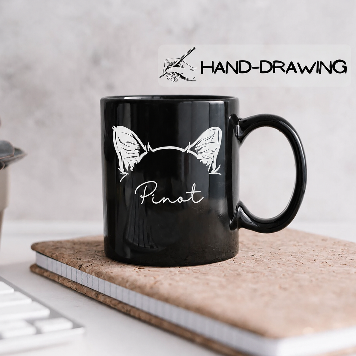 A glossy black ceramic mug with a white hand-drawn style graphic of cat ears and the word 'Pinot' sits on a cork notebook on a desk. A white banner overlay at the top reads 'HAND-DRAWING' with a small hand-drawing icon.
