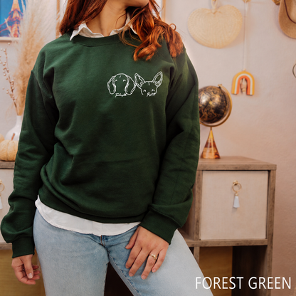 A woman wearing a forest green crewneck sweatshirt with a white line-art graphic of two French bulldogs on the chest, paired with light blue jeans in a cozy, decorated room.