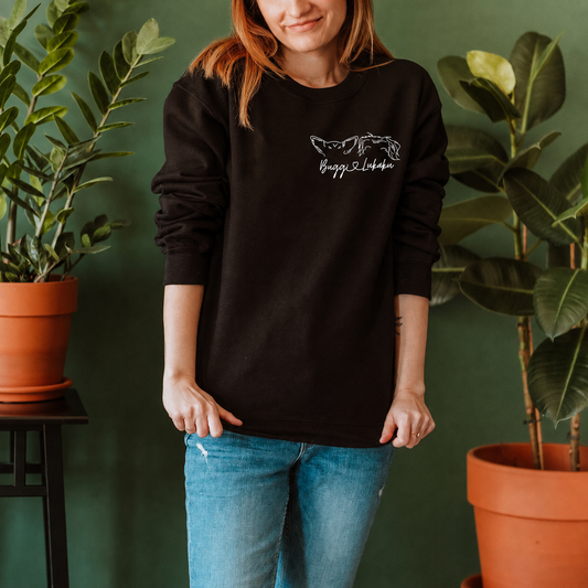 A woman wearing a black crewneck sweatshirt with a white graphic of a dog and the text 'Buddy & Lulu' on the chest, paired with blue jeans, standing in front of a dark green wall with potted plants.