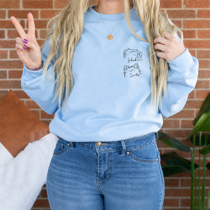 A woman wearing a light blue crewneck sweatshirt with a small black line-art graphic on the left chest, paired with blue jeans, posing in front of a brick wall background.