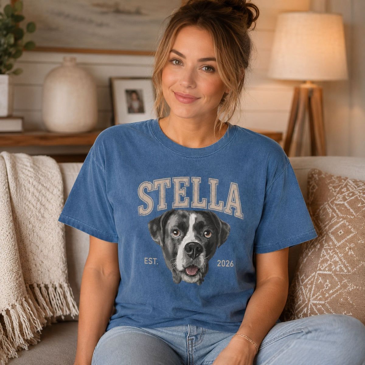 A woman sitting on a couch wearing a blue graphic t-shirt with a custom dog portrait and the name 'STELLA'.