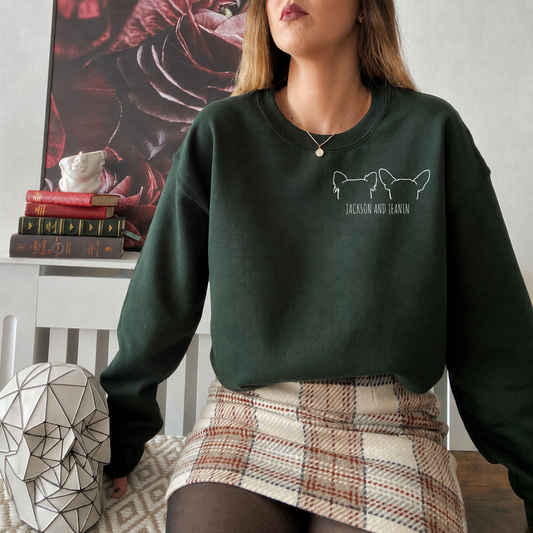 A woman wearing a dark forest green crewneck sweatshirt with a white graphic of two dogs and the text 'JACKSON AND BAYON' on the chest, paired with a plaid mini skirt in an indoor setting.