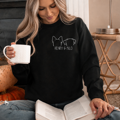 A woman sitting in front of a fireplace wearing a black sweatshirt with a white line art graphic of two dogs and the text 'HENRY & MISO'. She is holding a white mug and reading a book, with pumpkins on the floor nearby.