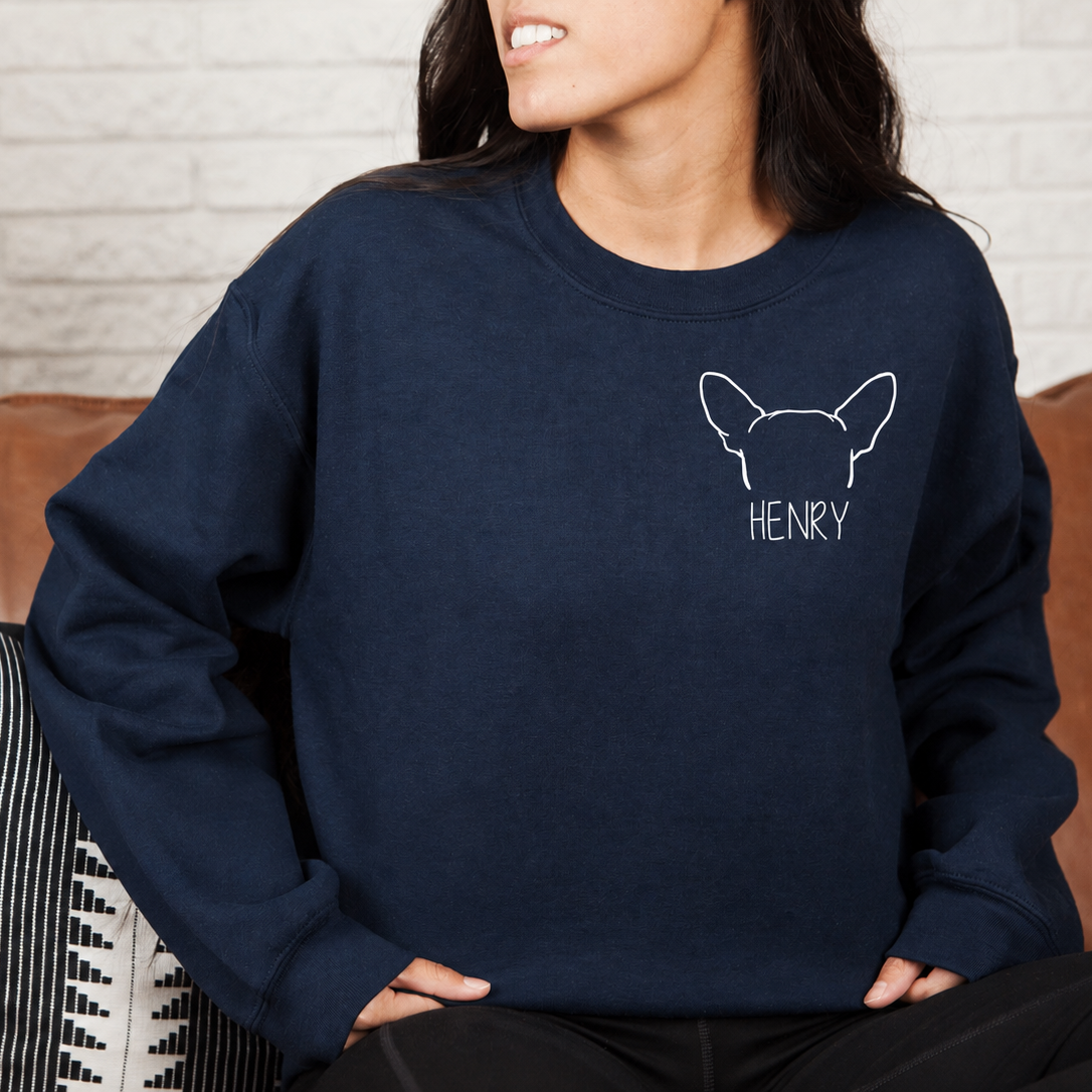 A woman wearing a navy blue crewneck sweatshirt with a white outline graphic of a French Bulldog and the name 'HENRY' printed on the chest, sitting on a brown leather couch.
