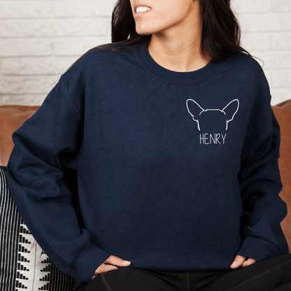 A woman wearing a navy blue crewneck sweatshirt with a white outline graphic of a French Bulldog and the name 'HENRY' printed on the chest, sitting on a brown leather couch.