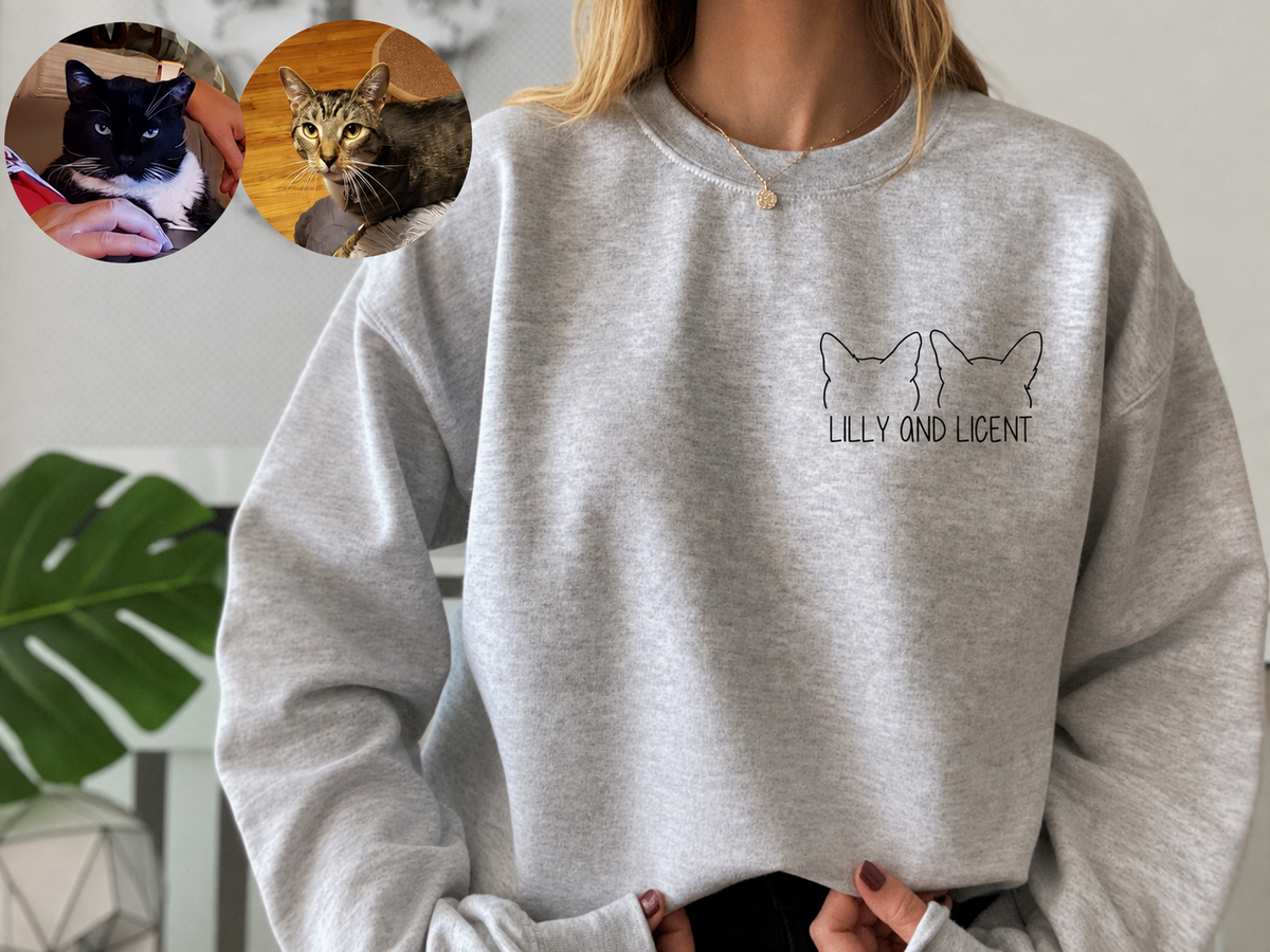 A person wearing a light heather gray crewneck sweatshirt with a black line-art graphic of two cat heads and the text 'LILLY AND LICENT' on the chest. Two circular inset photos in the top-left show different cats.