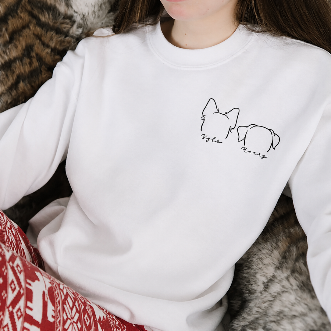 A person wearing a white crewneck sweatshirt with a small black line art graphic of a cat and dog on the left chest, paired with red and white patterned knit pants, sitting on a gray faux fur blanket.