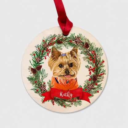 Round personalized pet ornament showing a small tan dog in a holiday wreath with a red name ribbon