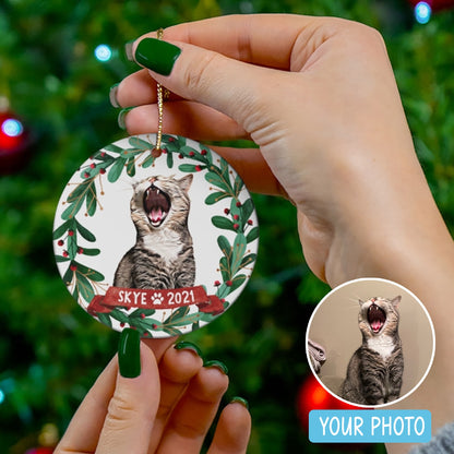 Hand holding a personalized cat ornament with leafy wreath, red ribbon, and name Skye