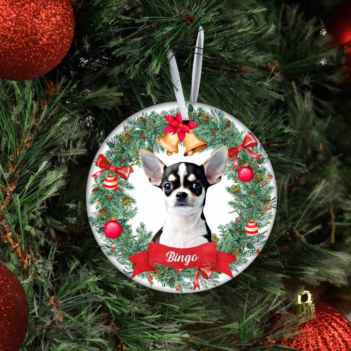 A personalized Christmas ornament featuring a Chihuahua dog portrait inside a green wreath with red bows and gold bells, hanging on a Christmas tree with red ornaments.