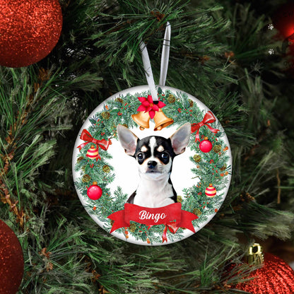 A personalized Christmas ornament featuring a Chihuahua dog portrait inside a green wreath with red bows and gold bells, hanging on a Christmas tree with red ornaments.