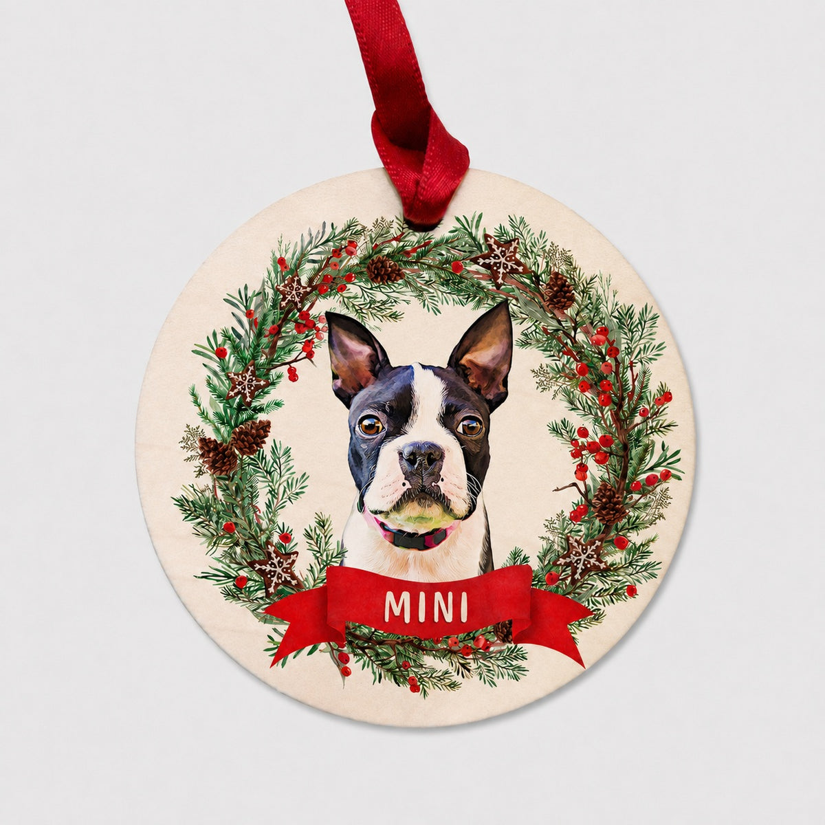 Round personalized pet ornament showing a Boston Terrier in a festive evergreen wreath with a red name ribbon