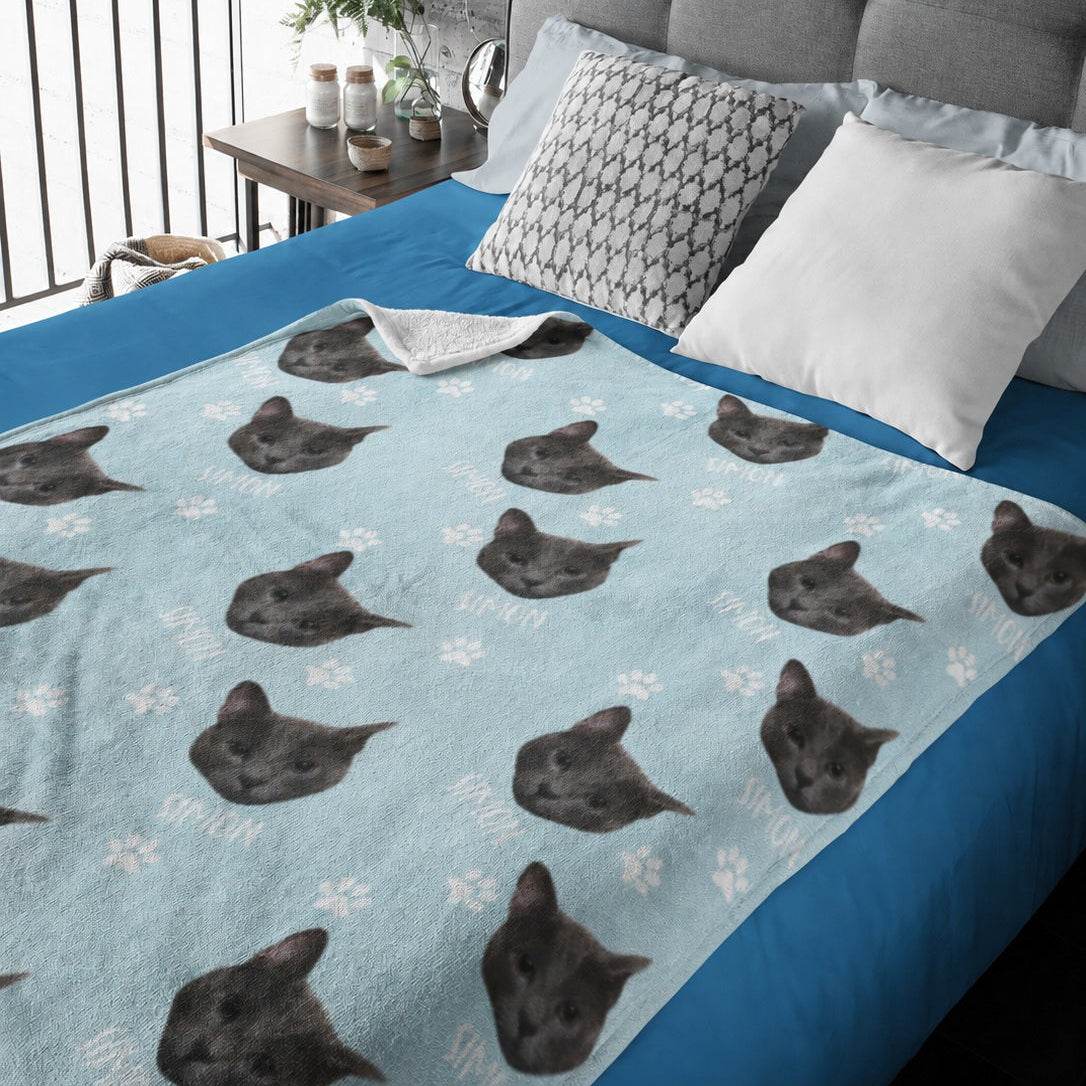 Light blue custom pet blanket with repeated dark cat portraits, white name text, and paw prints on a bed