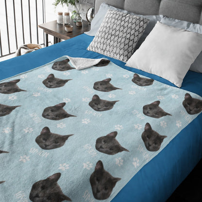 Light blue custom pet blanket with repeated dark cat portraits, white name text, and paw prints on a bed