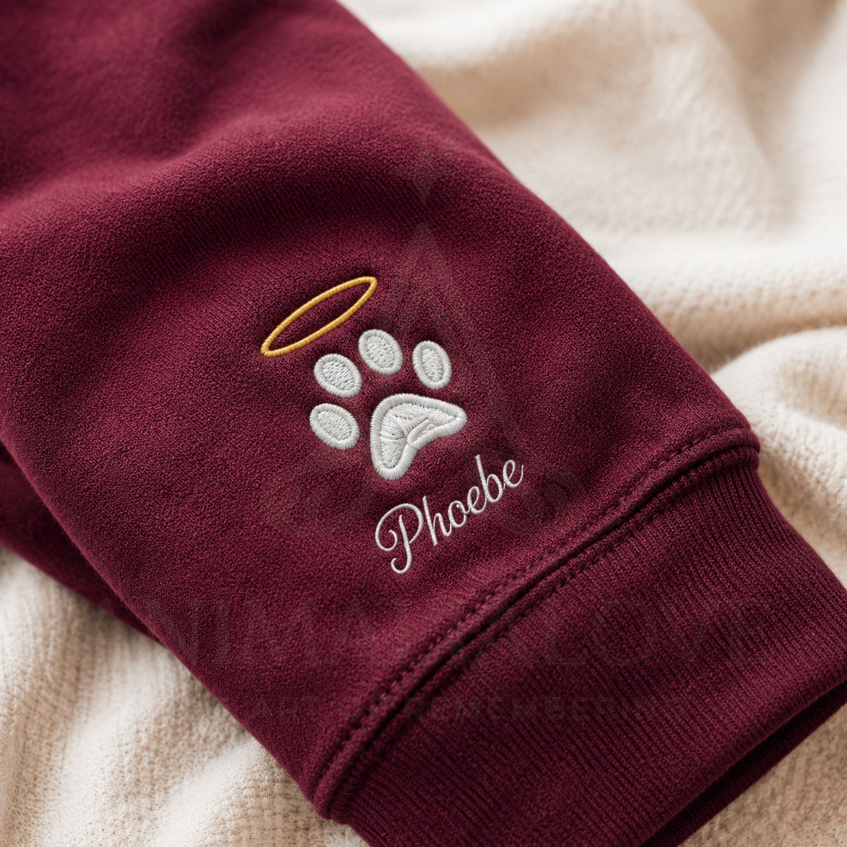 A close-up of a burgundy fleece sweatshirt sleeve with an embroidered white paw print, gold halo, and the name 'Phoebe' in white script, resting on a cream sherpa fabric.