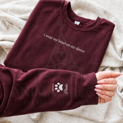 A folded maroon sweatshirt laid on a soft beige blanket, with a hand holding the cuff. The sweatshirt has white text reading 'I wear my heart on my sleeve' and a small paw print graphic with the name 'Phebe' on the cuff.
