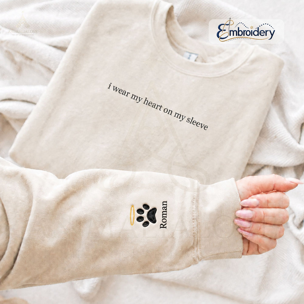 A beige crewneck sweatshirt laid flat on a matching beige blanket, with a person's hand holding up the sleeve to show a custom embroidery design. The sleeve features a black paw print with a small gold halo and the name 'Roman' in black text.