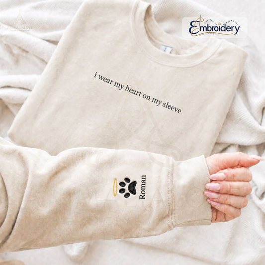 A beige crewneck sweatshirt laid flat on a matching beige blanket, with a person's hand holding up the sleeve to show a custom embroidery design. The sleeve features a black paw print with a small gold halo and the name 'Roman' in black text.
