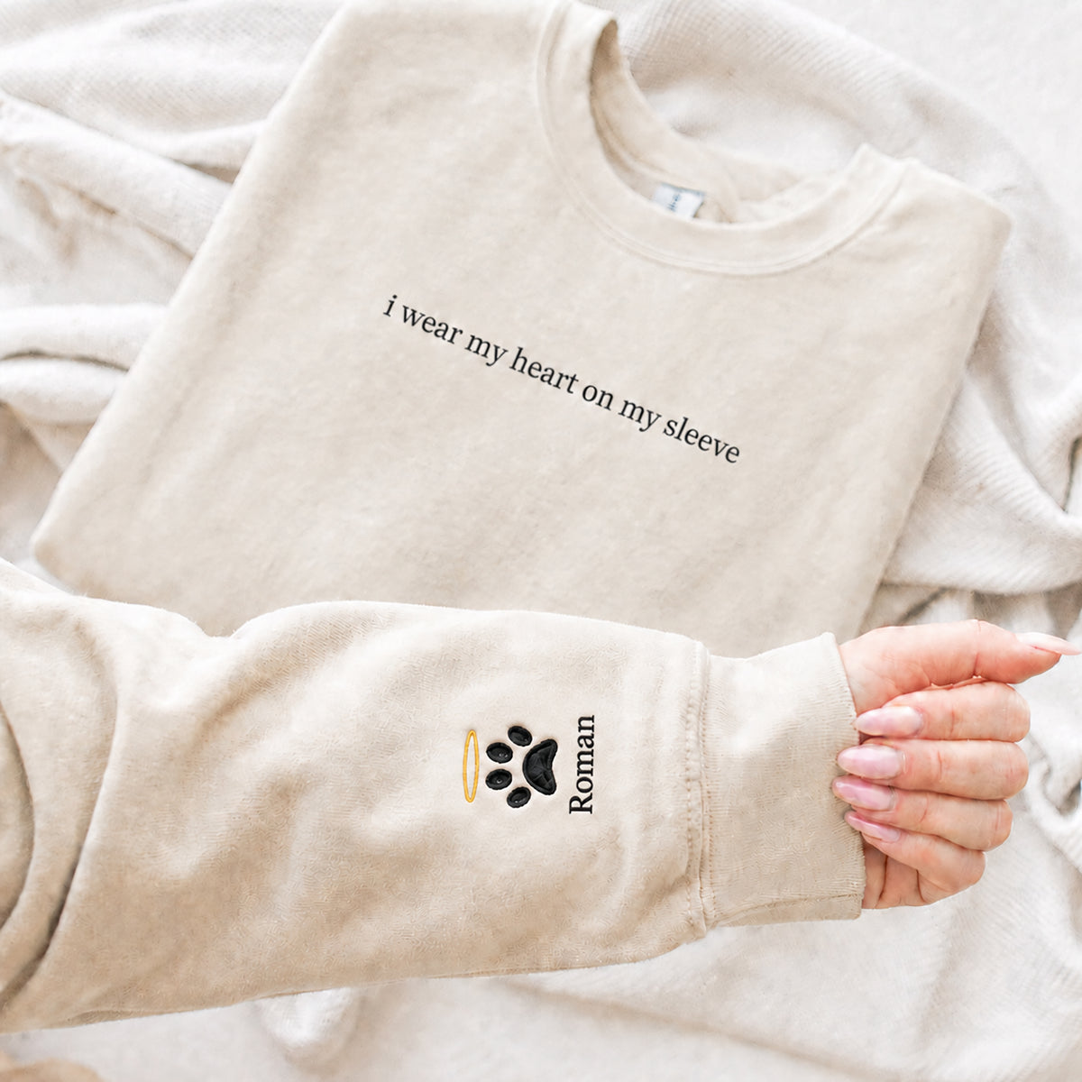 A beige crewneck sweatshirt laid flat on a soft cream blanket, with a person's hand holding up the sleeve to show a custom black paw print and the name 'Roman' alongside a small gold halo.