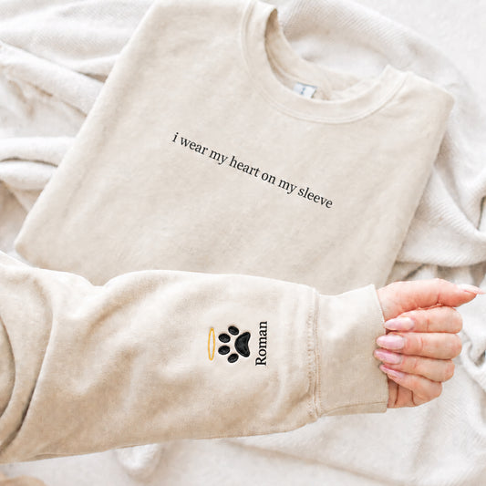 A beige crewneck sweatshirt laid flat on a soft cream blanket, with a person's hand holding up the sleeve to show a custom black paw print and the name 'Roman' alongside a small gold halo.