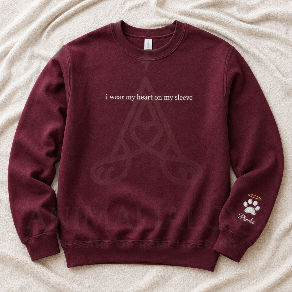 A maroon crewneck sweatshirt laid flat on a beige fabric background. The front has white text reading 'i wear my heart on my sleeve' above a faint Celtic knot design, and the left sleeve has a small white paw print with a halo and the word 'Peano'.