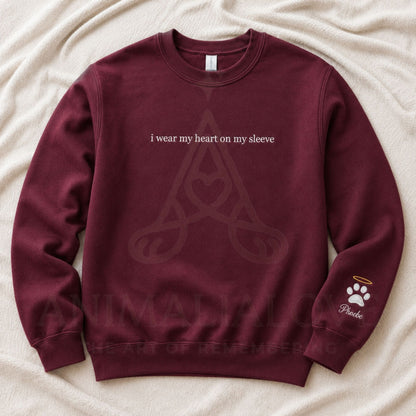 A maroon crewneck sweatshirt laid flat on a beige fabric background. The front has white text reading 'i wear my heart on my sleeve' above a faint Celtic knot design, and the left sleeve has a small white paw print with a halo and the word 'Peano'.