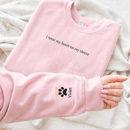 A folded light pink sweatshirt laid on a soft cream fabric background, with one sleeve extended across the front. The sweatshirt has black text reading 'I wear my heart on my sleeve' across the chest and a small black paw print with a gold ring and the name 'Violet' on the cuff.