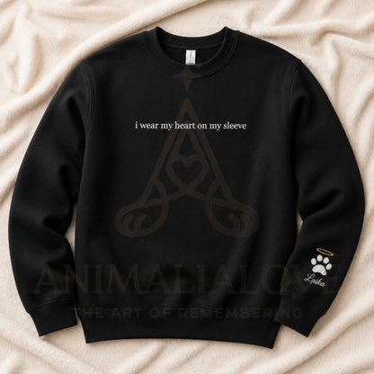 A black crewneck sweatshirt laid flat on a beige fabric background. The front has white text reading 'i wear my heart on my sleeve' above a dark gray Celtic knot design with a heart.