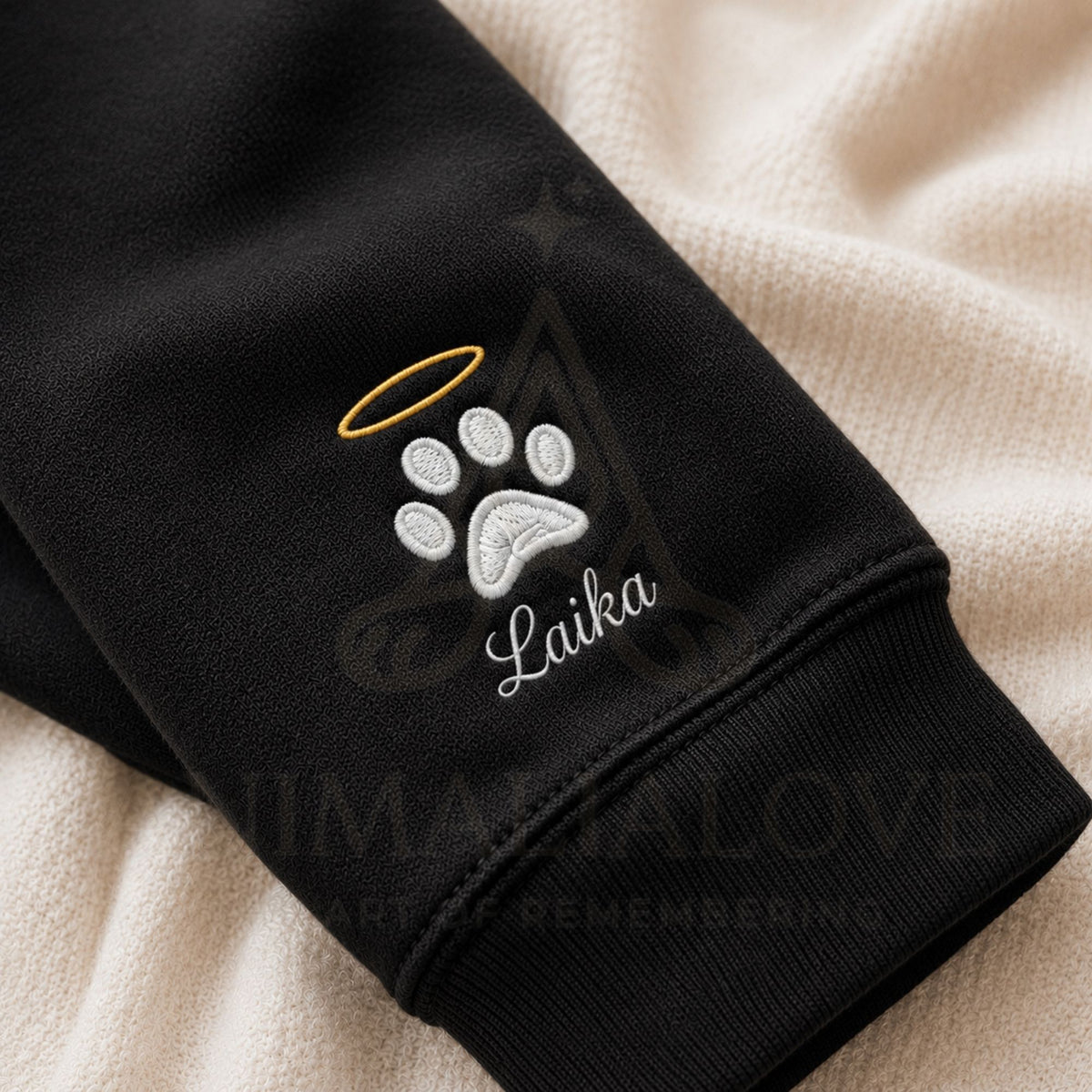 Close-up of a black sweatshirt cuff featuring an embroidered white paw print with a gold halo and the name 'Laika' in white script, laid over a cream textured blanket. A faint 'HUMANA LOVE' watermark is visible across the image.