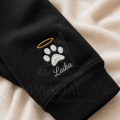 Close-up of a black sweatshirt cuff featuring an embroidered white paw print with a gold halo and the name 'Laika' in white script, laid over a cream textured blanket. A faint 'HUMANA LOVE' watermark is visible across the image.